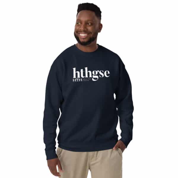 A smiling man with short curly hair and a beard wears a unisex crew neck sweatshirt featuring a bold white logo, along with smaller text below, and stands with one hand in his pocket against a plain background.