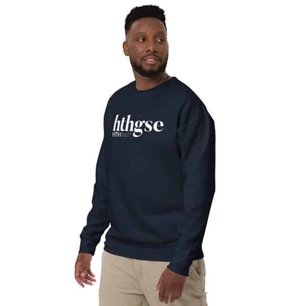 A man in a navy blue Unisex Crew Neck Sweatshirt with a white logo stands against a white background, looking slightly to his right. He is also wearing light-colored pants.