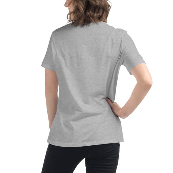 A person with wavy brown hair, wearing a plain light gray Women's Relaxed Fit T-Shirt and black pants, is shown from the back with one hand on their hip.