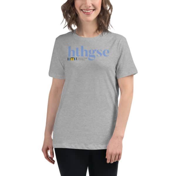 A smiling woman with wavy brown hair wears a light gray Women's Relaxed Fit T-Shirt with blue text hthgse and a small black and yellow HTH Graduate School of Education logo beneath it. She stands against a plain white background.