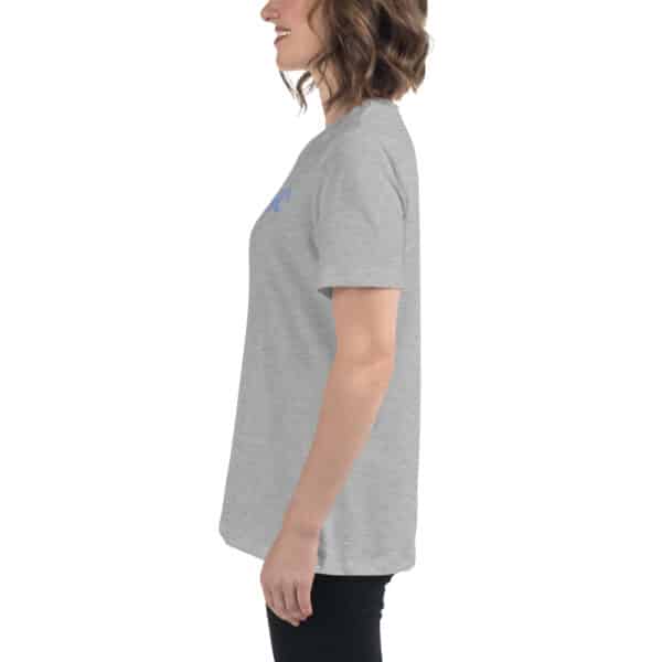 A woman with wavy brown hair is smiling and standing in profile, wearing a women's relaxed fit t-shirt in light gray and black pants against a plain white background.
