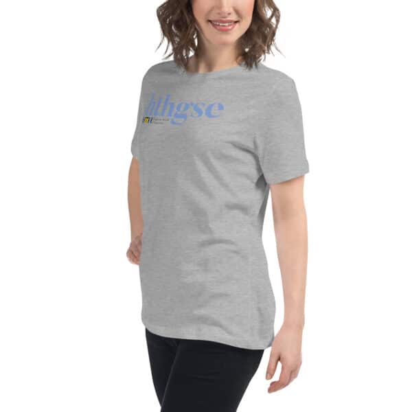 A woman with wavy brown hair smiles while wearing a Women's Relaxed Fit T-Shirt in light gray with blue and yellow text across the chest. She stands against a white background, facing slightly to the side.