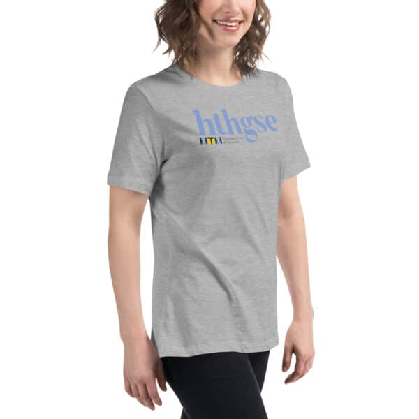 A woman with wavy brown hair wears a heather gray Women's Relaxed Fit T-Shirt featuring large blue text hthgse and smaller multicolored text below. She is smiling and standing against a white background.