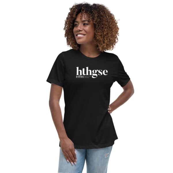 A smiling woman with curly hair wears a Women's Relaxed Fit T-Shirt in black with “hthgse” printed in white. She stands confidently with one hand on her hip, pairing the T-shirt with light blue jeans.