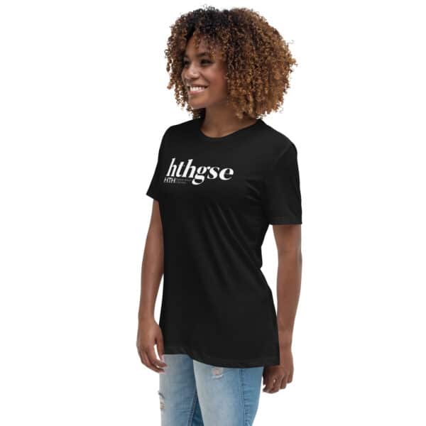 A woman with curly hair wears a Women's Relaxed Fit T-Shirt in black, featuring a bold white logo that reads hthgse. She smiles and looks to the side, paired with light blue ripped jeans against a plain white background.
