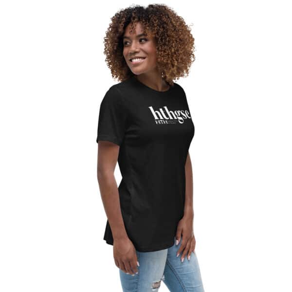 A woman with curly hair smiles and looks to the side while wearing a black Women's Relaxed Fit T-Shirt with hthgse printed on it, paired with light blue ripped jeans, standing against a white background.