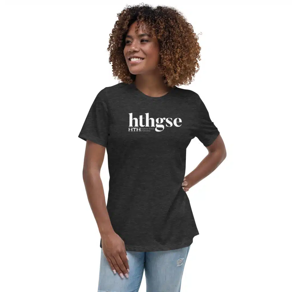 A smiling woman with curly hair wears a Women's Relaxed Fit T-Shirt in dark gray, featuring a bold white logo with the text hthgse, standing confidently with one hand on her hip against a plain white background.