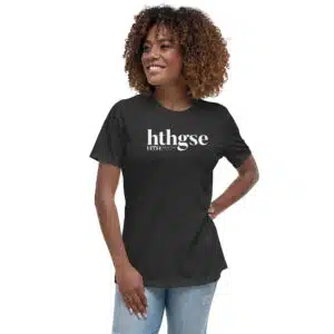 A smiling woman with curly hair wears a Women's Relaxed Fit T-Shirt in dark gray, featuring a bold white logo with the text hthgse, standing confidently with one hand on her hip against a plain white background.