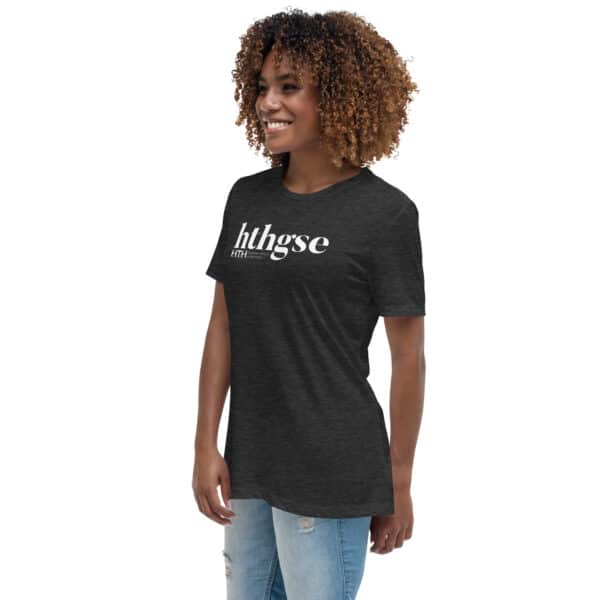 Smiling woman with curly hair wearing a Women’s Relaxed Fit T-Shirt in dark heather with "hthgse" printed in white, standing sideways and facing left, paired with light blue ripped jeans against a plain white background.