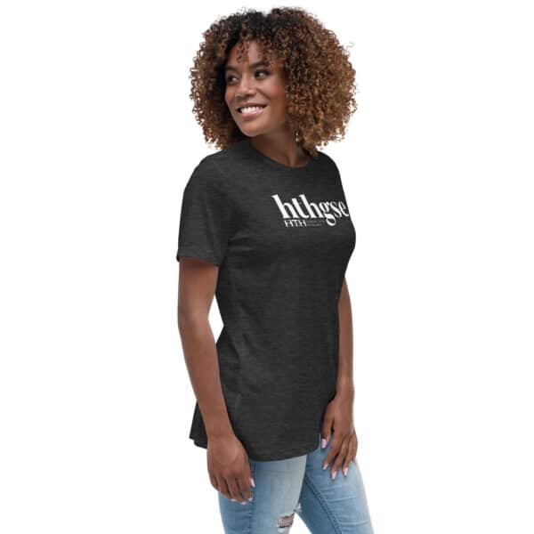 A woman with curly hair smiles while wearing a dark gray Women’s Relaxed Fit T-Shirt with hthgse printed in white text and light blue ripped jeans, standing against a plain white background.