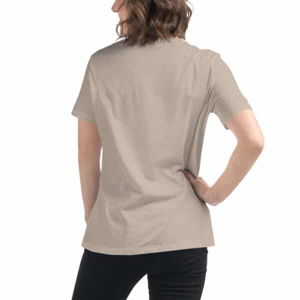 A person with wavy brown hair stands with their back to the camera, wearing a beige Women's Relaxed Fit T-Shirt and black pants. One hand rests on their hip while the other arm hangs by their side.