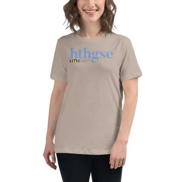 A smiling person with wavy brown hair wears a beige Women's Relaxed Fit T-Shirt featuring large blue text hthgse and a small HTH Graduate School of Education logo in black and yellow on the chest.