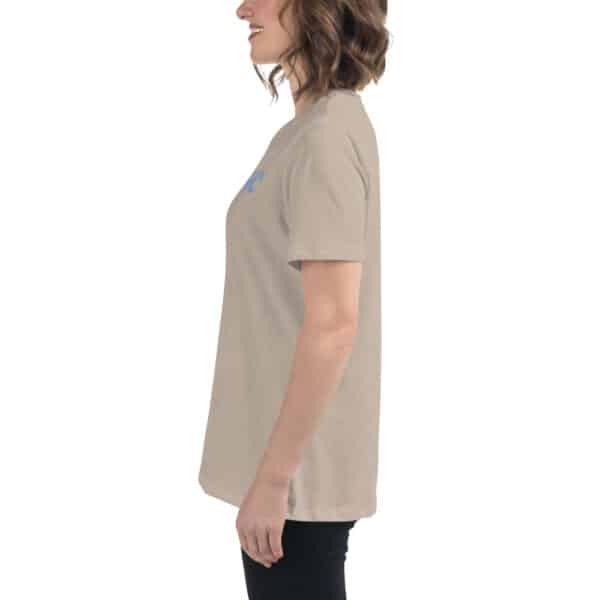 Woman standing in profile view, smiling with loose wavy hair, wearing a beige Women’s Relaxed Fit T-Shirt and black pants against a plain white background.