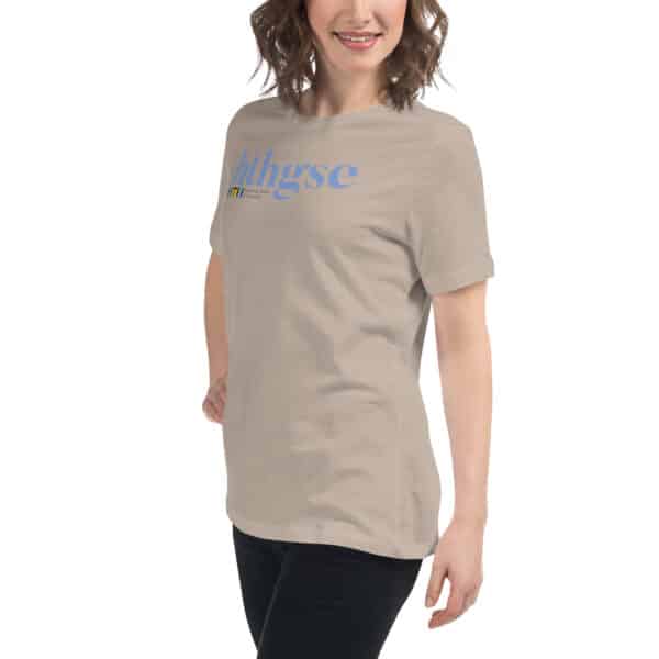 A woman with wavy brown hair smiles while wearing a Women's Relaxed Fit T-Shirt in beige, featuring blue "huhgse" text on the front. She is standing against a white background.