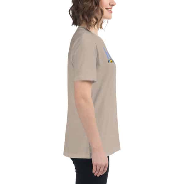 A woman with wavy brown hair is standing sideways, smiling in a full color Women's Relaxed Fit T-Shirt and black pants against a white background. The front of the women's t-shirt features a partially visible text or design.