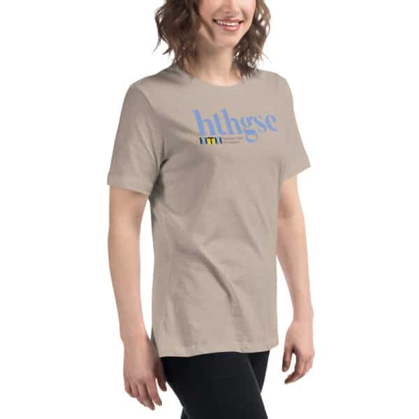 A smiling woman with wavy brown hair wears a light beige Women’s Relaxed Fit T-Shirt featuring blue “hthgse” text and a small colorful logo. She stands against a plain white background.