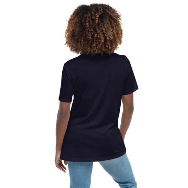 A woman with curly hair, seen from behind, wears a plain black Women's Relaxed Fit T-Shirt and light blue jeans. She stands with one hand in her pocket against a white background.