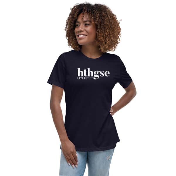 A smiling woman with curly hair wears a Women's Relaxed Fit T-Shirt featuring a white logo and bold text, standing with one hand on her hip against a plain white background.