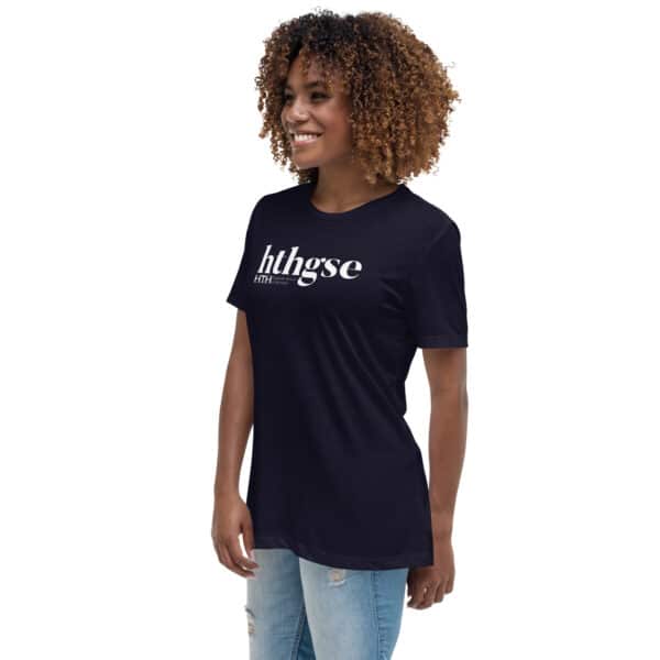 A person with curly hair smiles, wearing a Women's Relaxed Fit T-Shirt in black with "hthgse" in white, paired with light blue ripped jeans. The background is plain white.