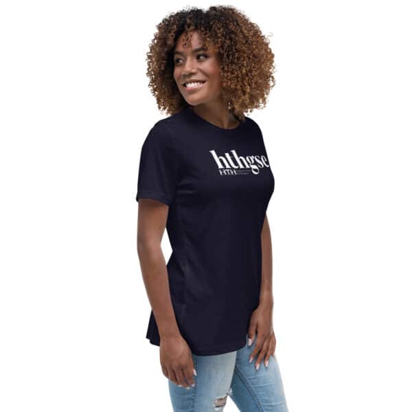 A woman with curly hair smiles while standing sideways, wearing a black Women’s Relaxed Fit T-Shirt that says htthgse in white text, paired with light blue ripped jeans against a white background.