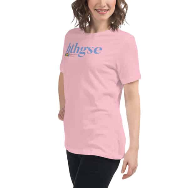 A woman wearing a Women's Relaxed Fit T-Shirt in light pink, featuring the letters hthgse in large blue text on the front, stands against a plain white background and smiles.