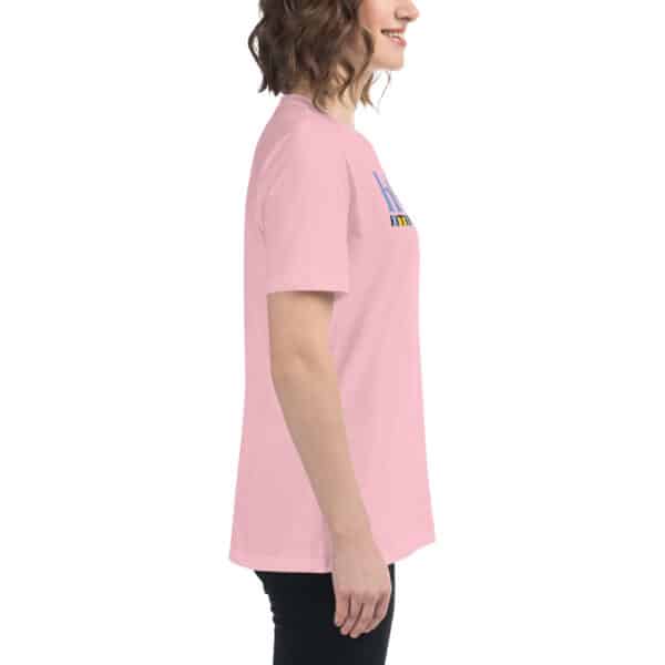 A woman stands in profile, smiling and wearing a Women's Relaxed Fit T-Shirt in light pink with graphic text, paired with black pants, against a plain white background.