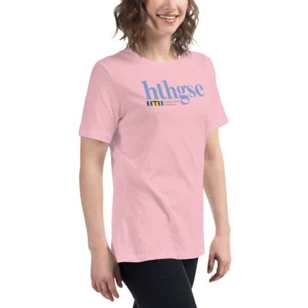 A smiling person with wavy brown hair wears a light pink Women's Relaxed Fit T-Shirt with blue text hthgse and a small colorful logo underneath on the upper front, standing against a plain white background.