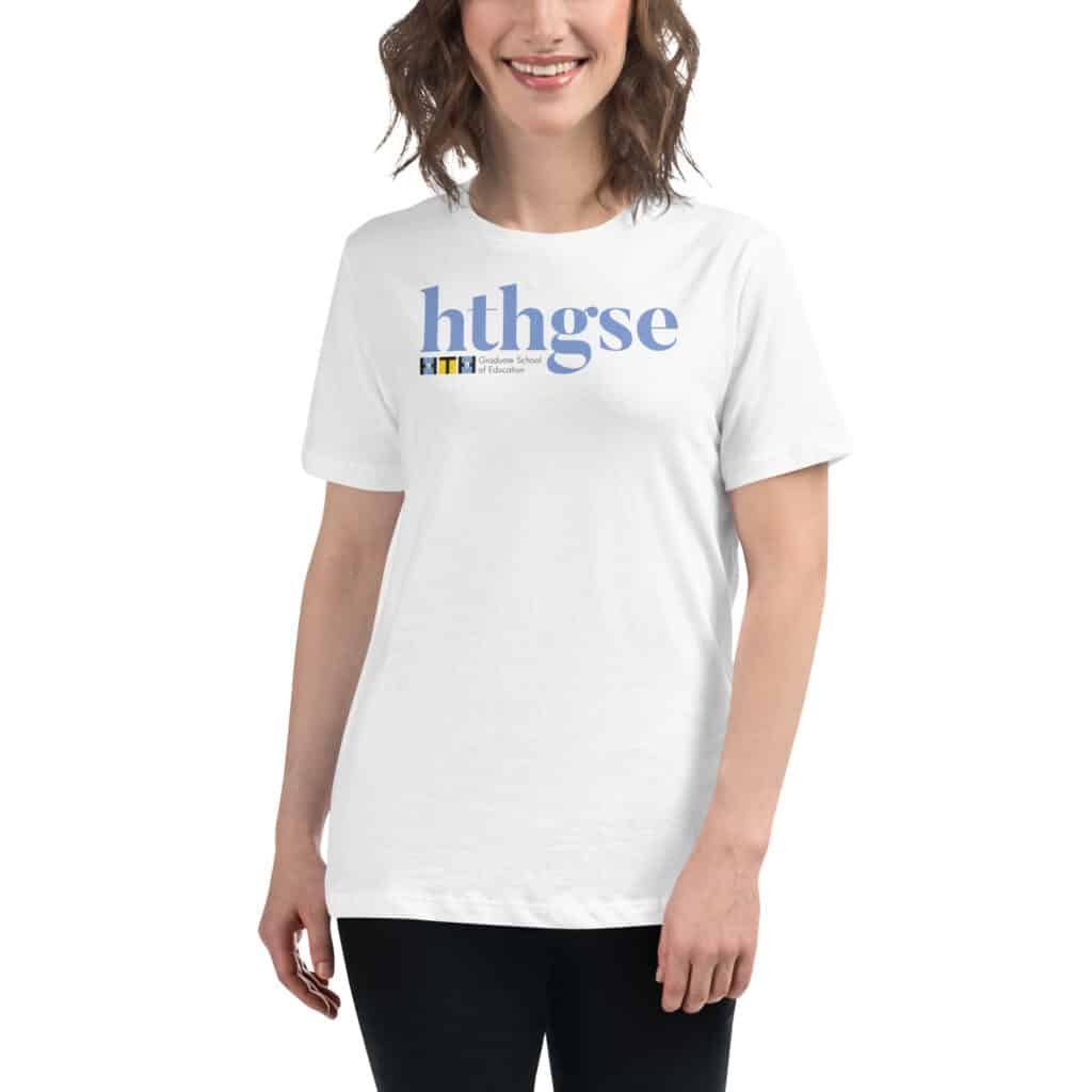 A woman with wavy brown hair is wearing a Women's Relaxed Fit T-Shirt that says hthgse in large blue letters with a small HTH Graduate School of Education logo underneath, standing and smiling.