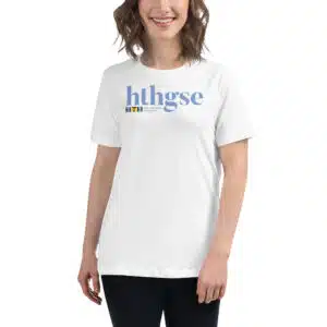 A woman with wavy brown hair is wearing a Women's Relaxed Fit T-Shirt that says hthgse in large blue letters with a small HTH Graduate School of Education logo underneath, standing and smiling.