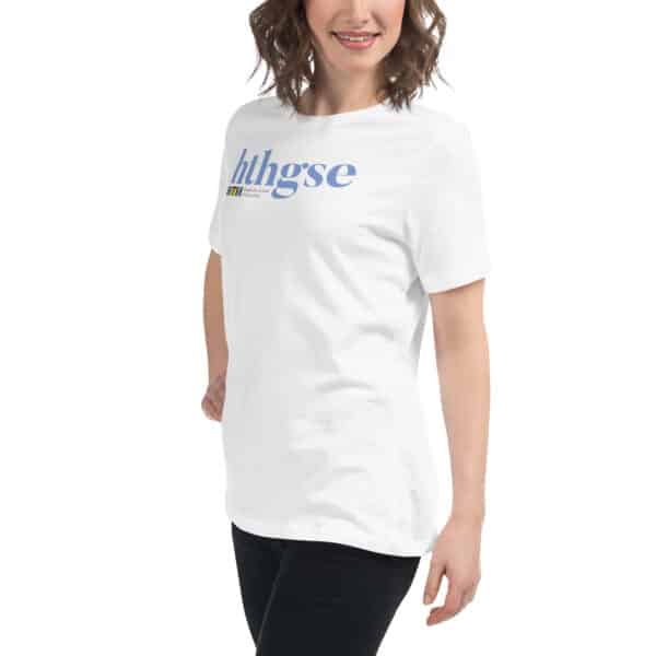 A woman with wavy brown hair is smiling and wearing a Women's Relaxed Fit T-Shirt with full color blue and yellow “hthgse” text. She stands against a plain white background.