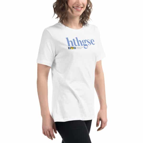 A smiling person with shoulder-length brown hair wears a Women's Relaxed Fit T-Shirt, featuring blue "hthgse" text and a colorful rectangular logo. The person stands against a plain white background.
