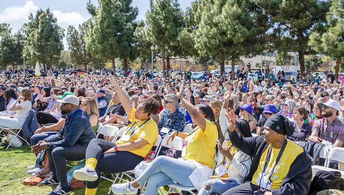 A large outdoor crowd sits on white chairs under tall trees, with many people raising their hands. The event appears lively and takes place on a sunny day in a park or grassy area.