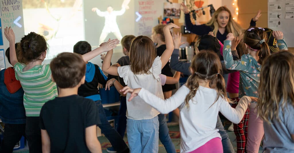 A group of young students stand in a colorful, lively classroom, raising their arms and moving together as they follow easy steps in a dance or exercise routine led by a teacher near a projected screen.