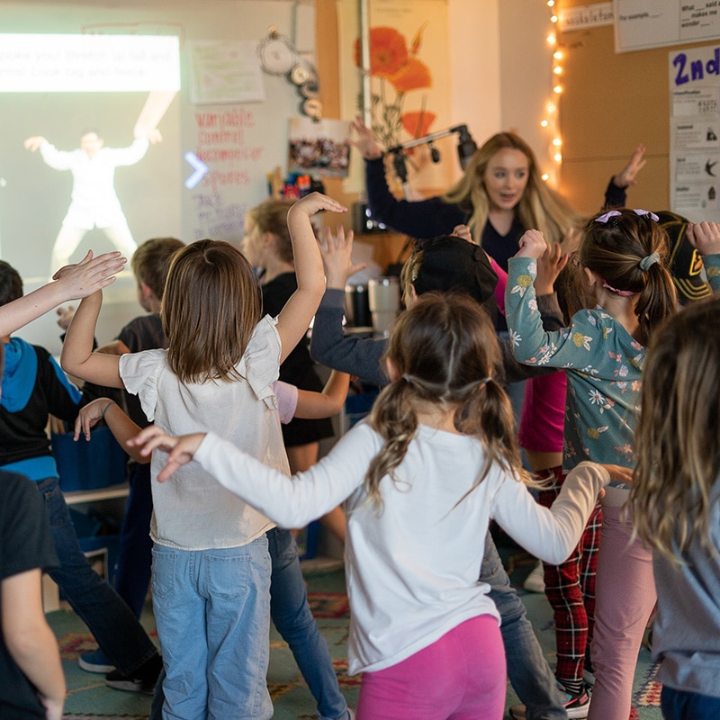 A group of young students are standing and raising their arms while following easy steps from a dance or exercise video projected on a classroom screen, with a female teacher guiding them in line with classroom norms.