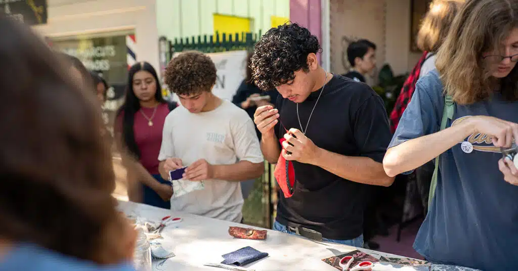 A group of young people stand around a table, focused on a sewing project. Scissors and sewing materials are spread out as they work together, learning new techniques during this creative lesson with fabric and thread.