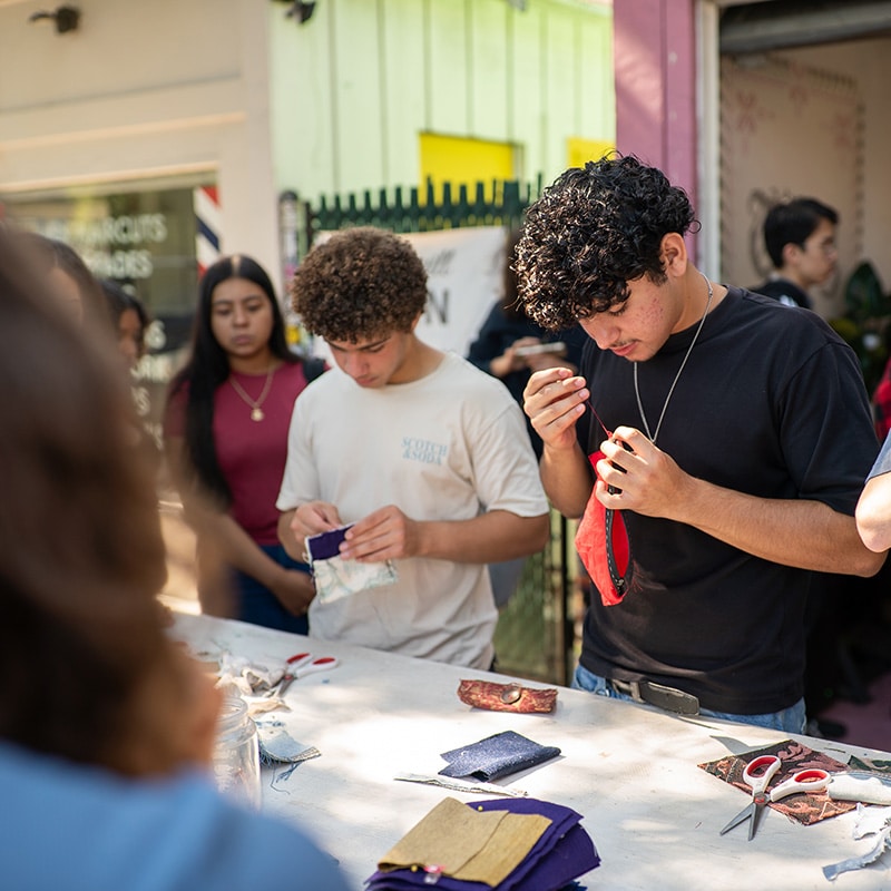 A group of young people stands around a table outdoors, focused on a fabric project for their unit, using scissors and thread. One person is threading a needle, while others work intently with their materials.