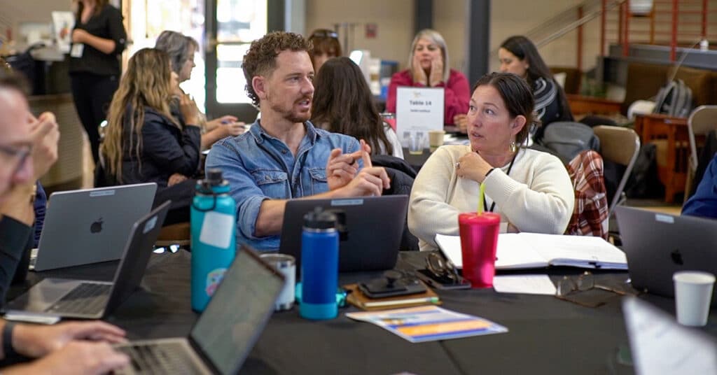 A group of people sit around a table with laptops, notebooks, and water bottles, engaged in a Dilemma Consultancy discussion. Two people in the middle are talking and gesturing, while others work or listen in the background.