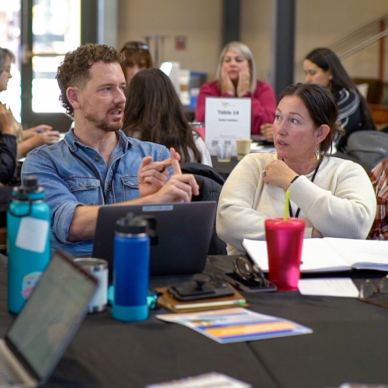 Two people sit at a table covered with laptops, notebooks, and water bottles, engaged in conversation during a Dilemma Consultancy workshop. Other participants are visible in the background.