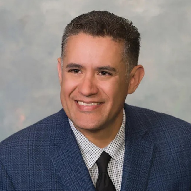 Moisés Aguirre, a man with short dark hair, wears a blue checkered suit, white patterned shirt, and black tie, smiling as he poses against a light gray background.