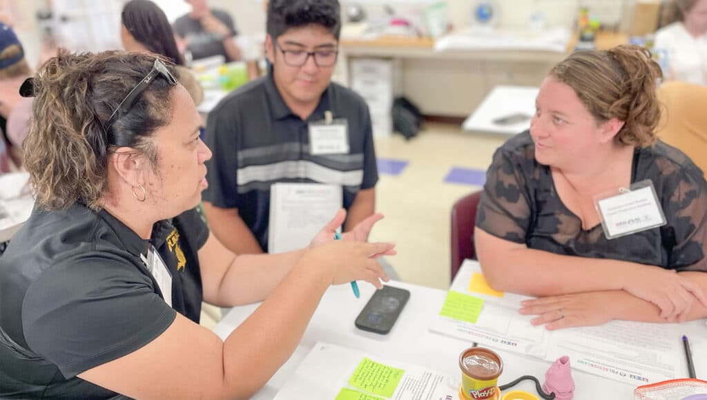 Three people sit at a table engaged in conversation, holding papers and pens. They appear to be in a classroom or office setting at PBL Design Camp, with documents, folders, and office supplies on the table.
