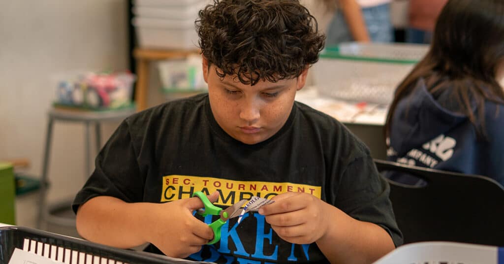 A young boy with curly hair concentrates as he uses green scissors to cut paper while sitting at a table in a productive classroom. Other students are visible in the background.