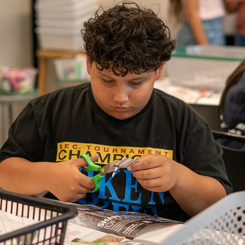 A young boy with curly hair sits at a table in a calm classroom, focused on cutting paper with green-handled scissors. He is wearing a black t-shirt with colorful text, surrounded by classroom materials and baskets.