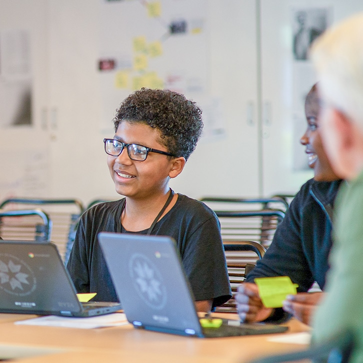 A smiling boy wearing glasses sits at a table with a laptop, holding a green sticky note. Another student beside him smiles, also holding a sticky note, as they engage in Math Curriculum activities in a classroom setting.
