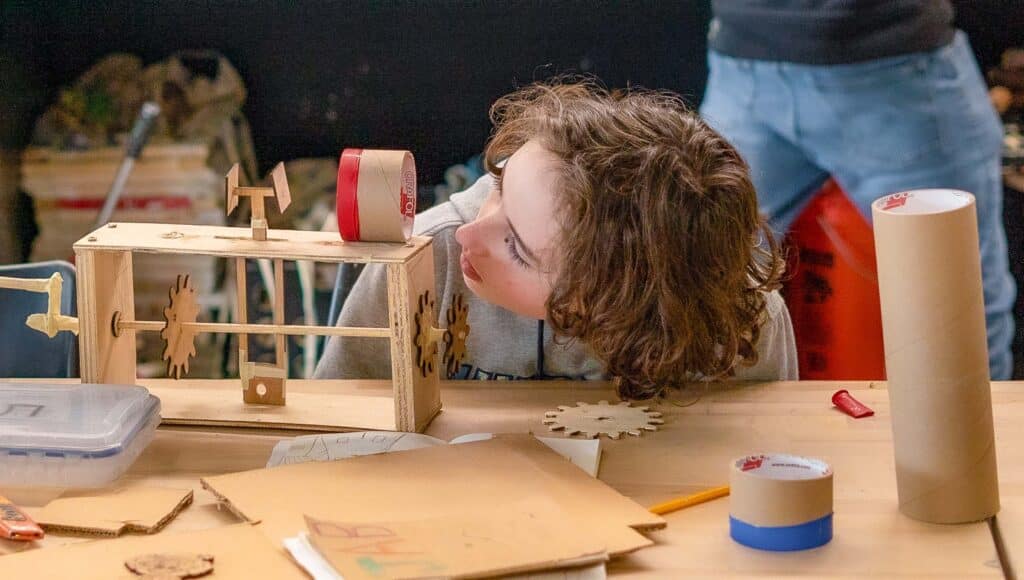 A child closely examines a wooden mechanical model on a cluttered table with papers, cardboard, tape, and art supplies, appearing focused and curious as they begin their Project Unpacking journey.