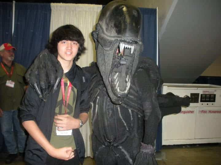 A teenager poses and smiles next to a person in a detailed, menacing Xenomorph costume from the Alien movie franchise at what appears to be an Imagine Mural Project convention. The Xenomorph has its arm around the teenager.