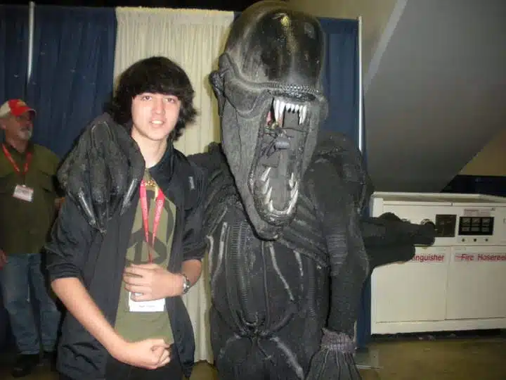 A teenager poses and smiles next to a person in a detailed, menacing Xenomorph costume from the Alien movie franchise at what appears to be an Imagine Mural Project convention. The Xenomorph has its arm around the teenager.