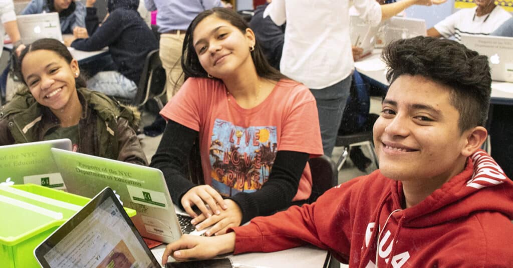 Three students sit at a classroom table, smiling at the camera. Each has a laptop in front of them, reflecting a school mindset that supports newcomer student integration as other students work together in the background.