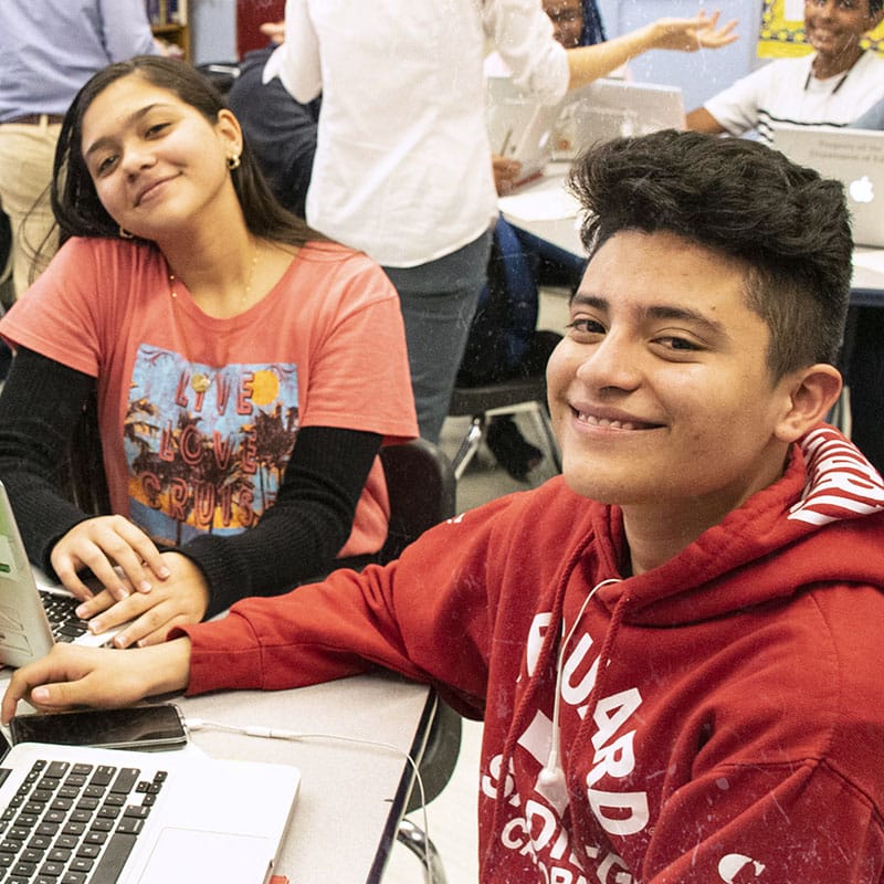 Two smiling teenagers sit at a table in a school classroom, working on laptops. The girl wears a red shirt over a black long-sleeve, and the boy wears a red hoodie. They appear focused while welcoming newcomer students into the group.