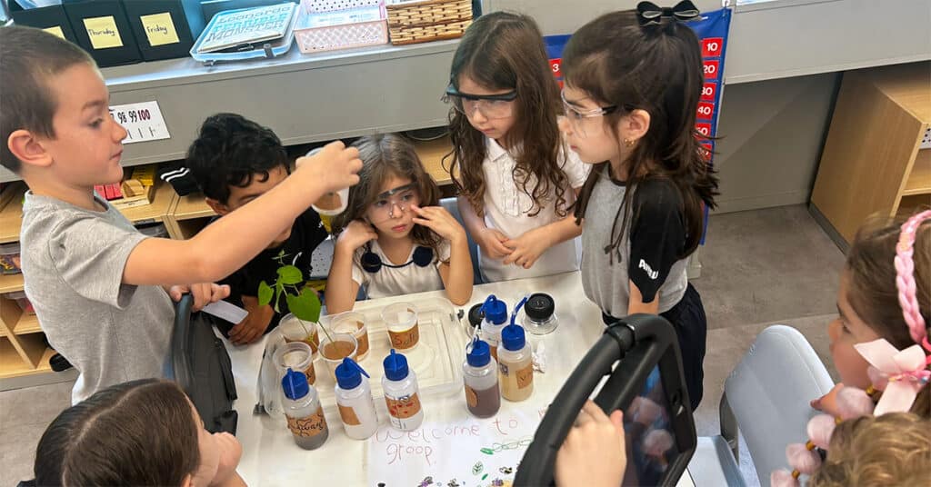 A group of first graders wearing safety glasses gather around a table with plants and bottles, observing a science experiment as part of a classroom community project.