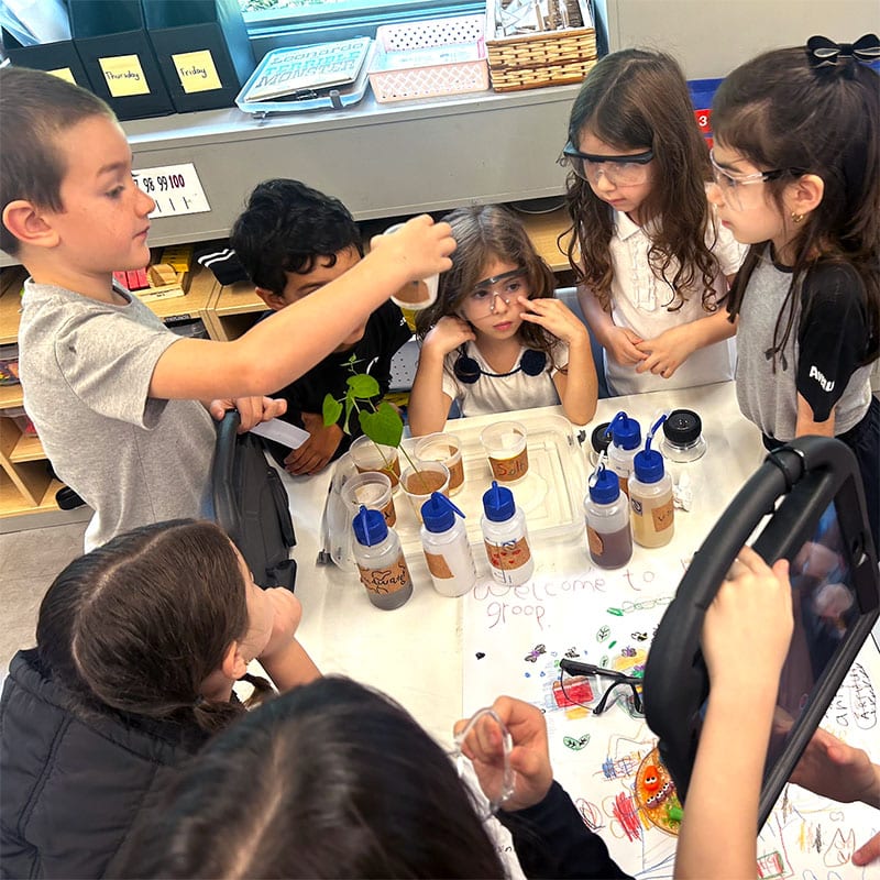 A group of first graders wearing safety glasses gathers around a table with various science experiment materials, observing as one child holds up a test tube with a plant inside during their co-designed project.
